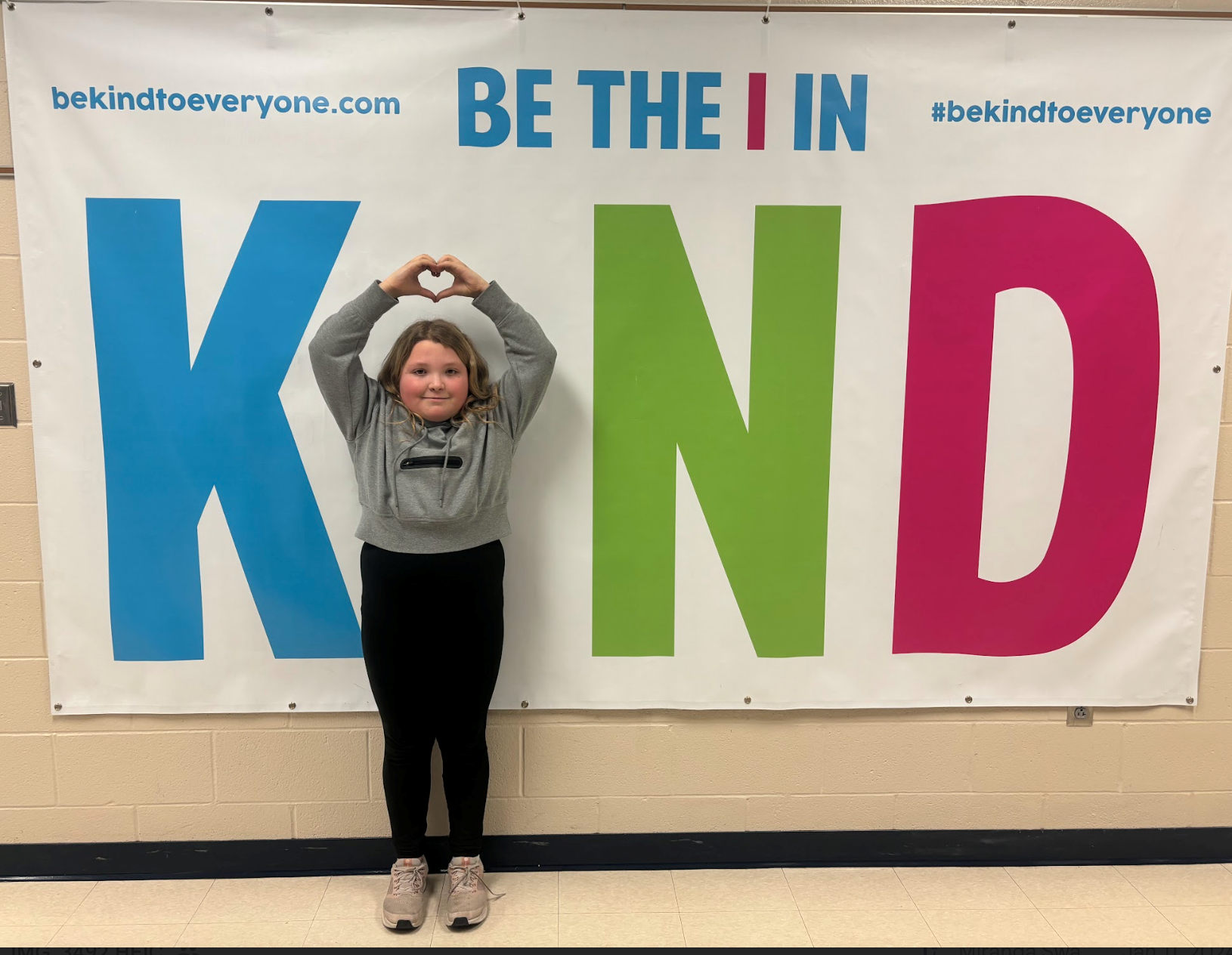 Student standing as the I making a heart above her head in a sign that says Be the I in KIND. 