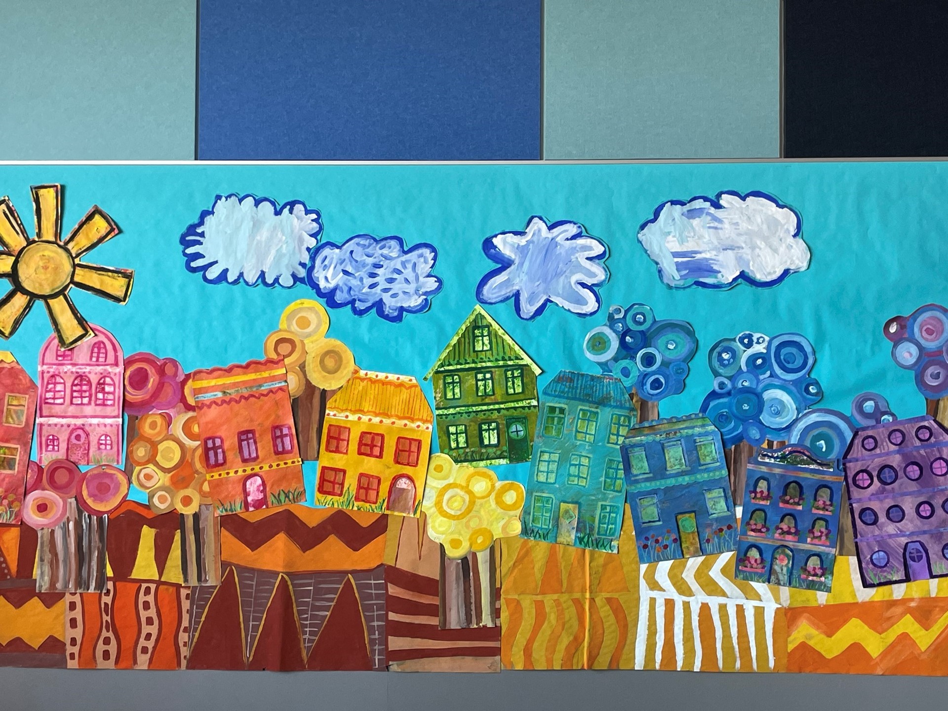 Student artwork; houses of different colors with trees, clouds and the sun. 